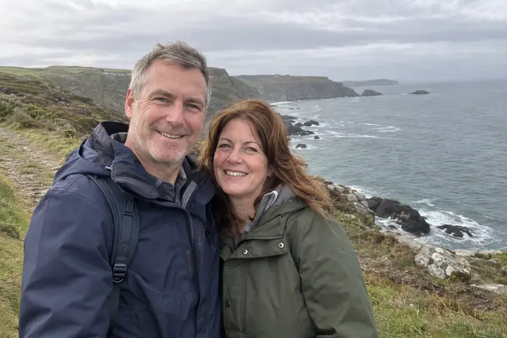 A couple in their fifties in rain jackets on a coastal clifftop path with the sea behind them, in color