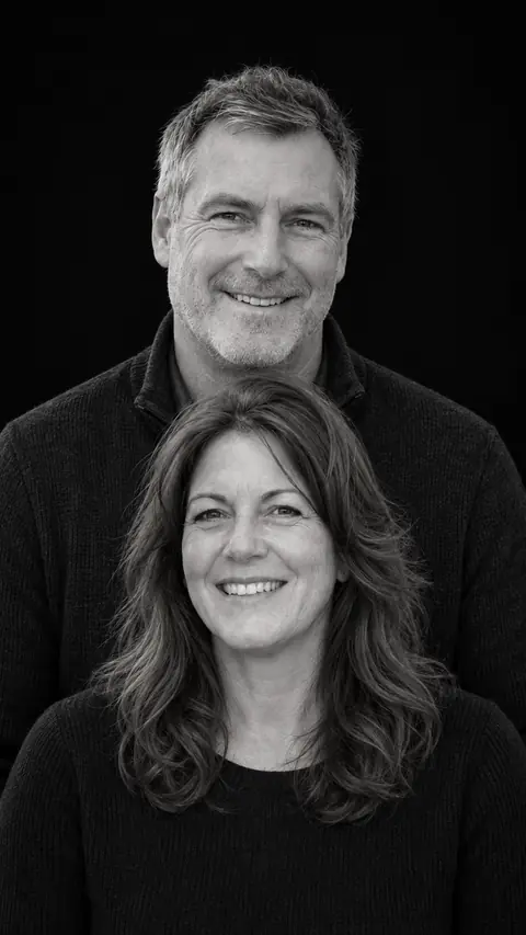 The same couple as a stacked monochrome studio portrait against black, the man behind the woman, both in dark sweaters