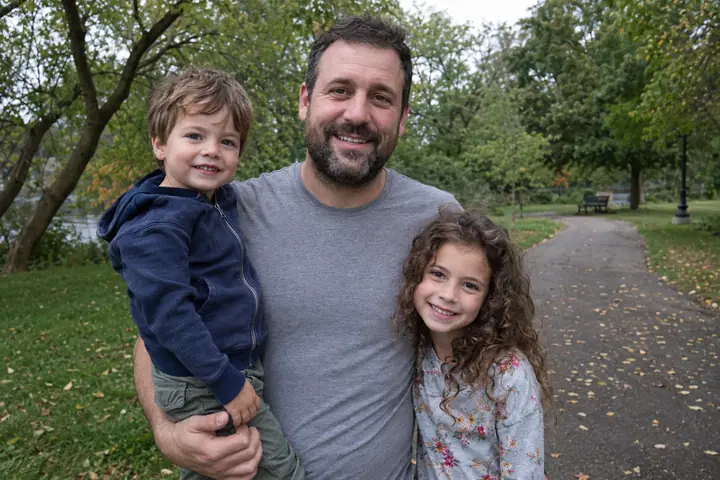 A father in a park holding his young son on one arm, his daughter with long curly hair standing beside him, all three smiling on an autumn afternoon in color