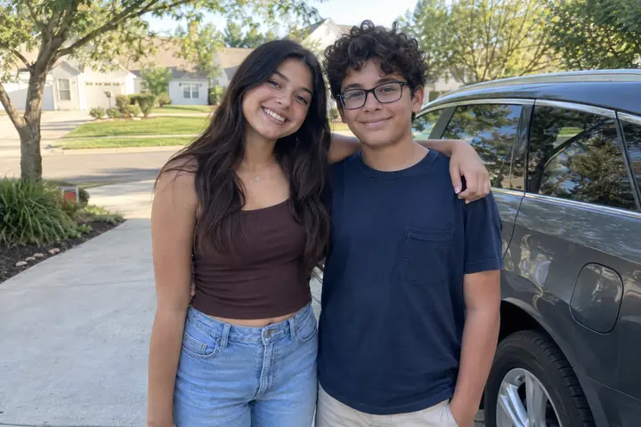 A teenage girl with long dark hair and a younger boy with curly hair and glasses, standing together outside on a suburban street in color