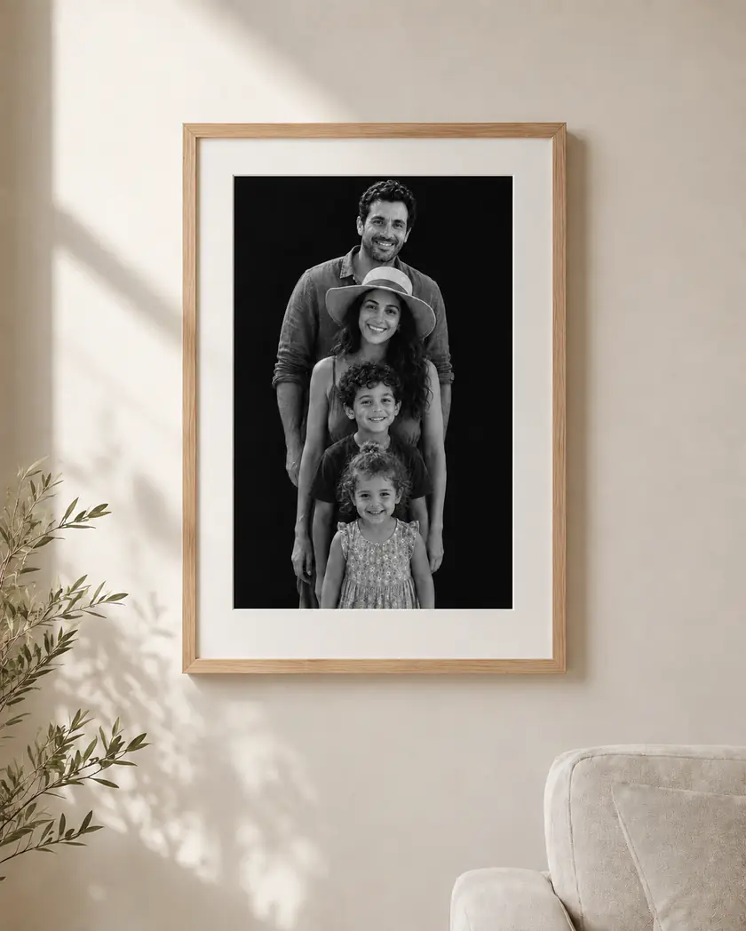 A stacked monochrome family portrait framed in light oak with a wide white mat, hanging on a warm plaster wall beside an olive branch in afternoon light